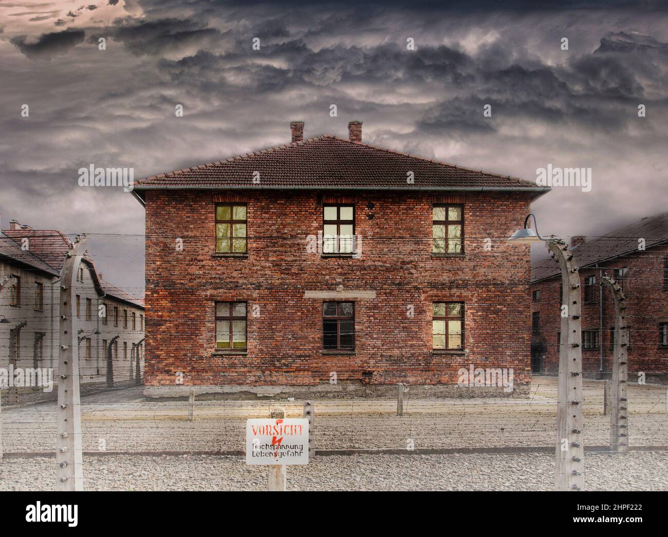 Casa in muratore dietro la recinzione di filo spinato elettrico e un tavolo con avvertimento in tedesco. Prigione dell'era del WW2. Foto Stock