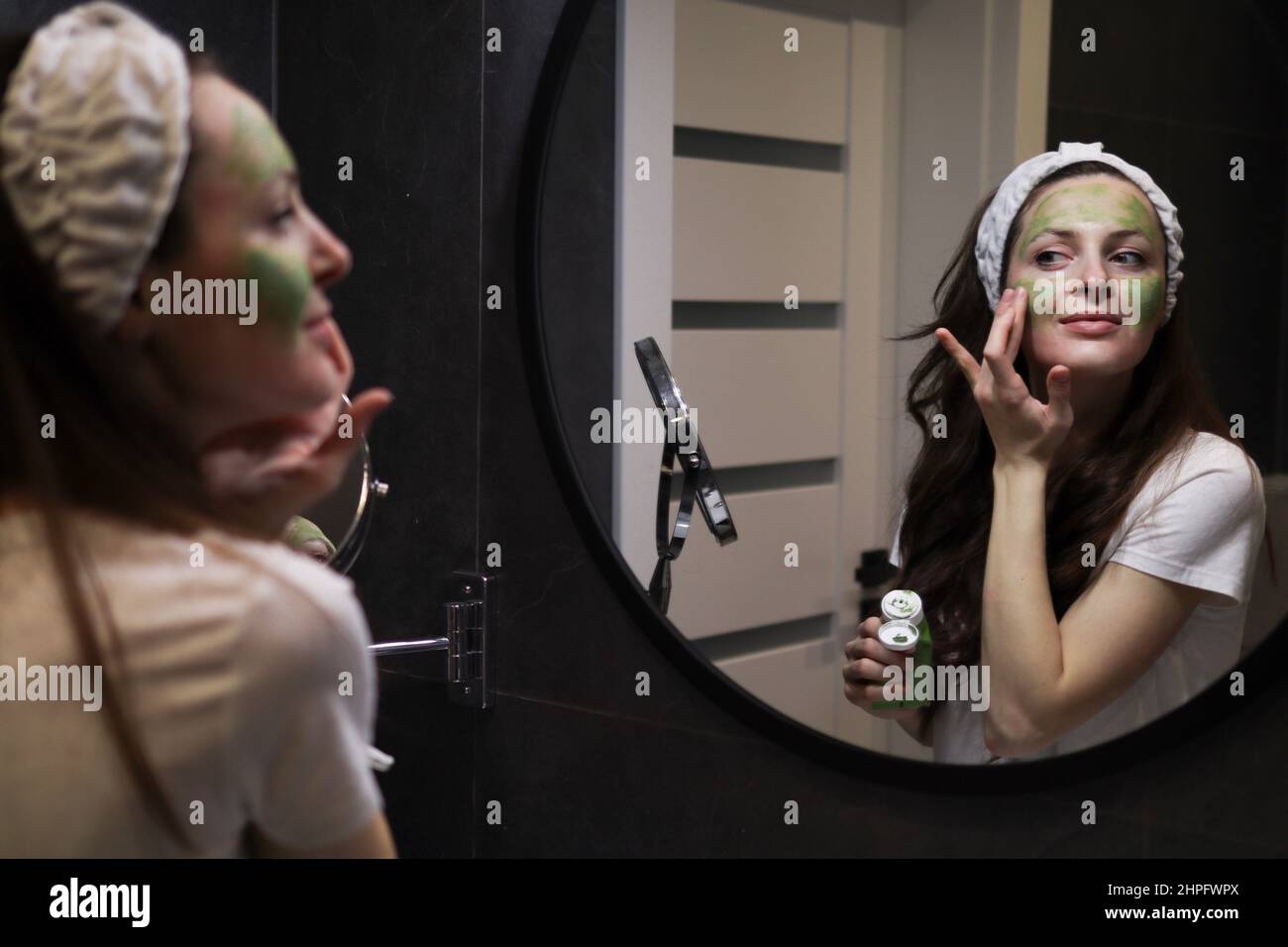 Donna in fascia applicando una maschera di argilla verde al suo viso in un bagno interno scuro di fronte a uno specchio. Ragazza che si prende cura di carnagione oleosa. Natura Foto Stock