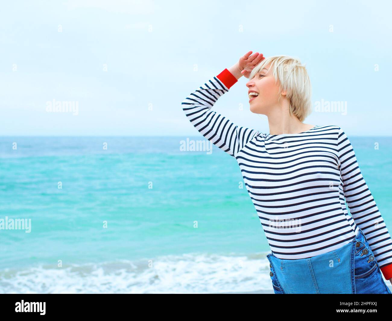 bella giovane bionda caucasica donna in vacanza in blusa a righe, sneakers e denim in generale sulla spiaggia, vicino al mare blu sorprendente sfondo. Foto Stock