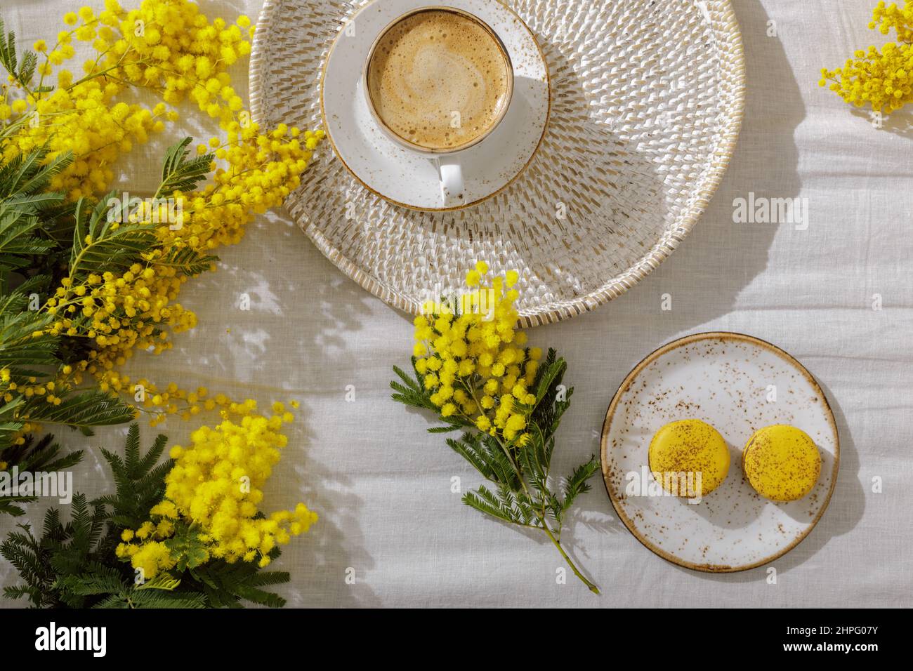 Rami di fiori gialli di mimosa su biancheria da letto bianca, tazza o caffè, amaretti gialli francesi. Saluto per la Giornata Internazionale delle Donne del 8t marzo Foto Stock