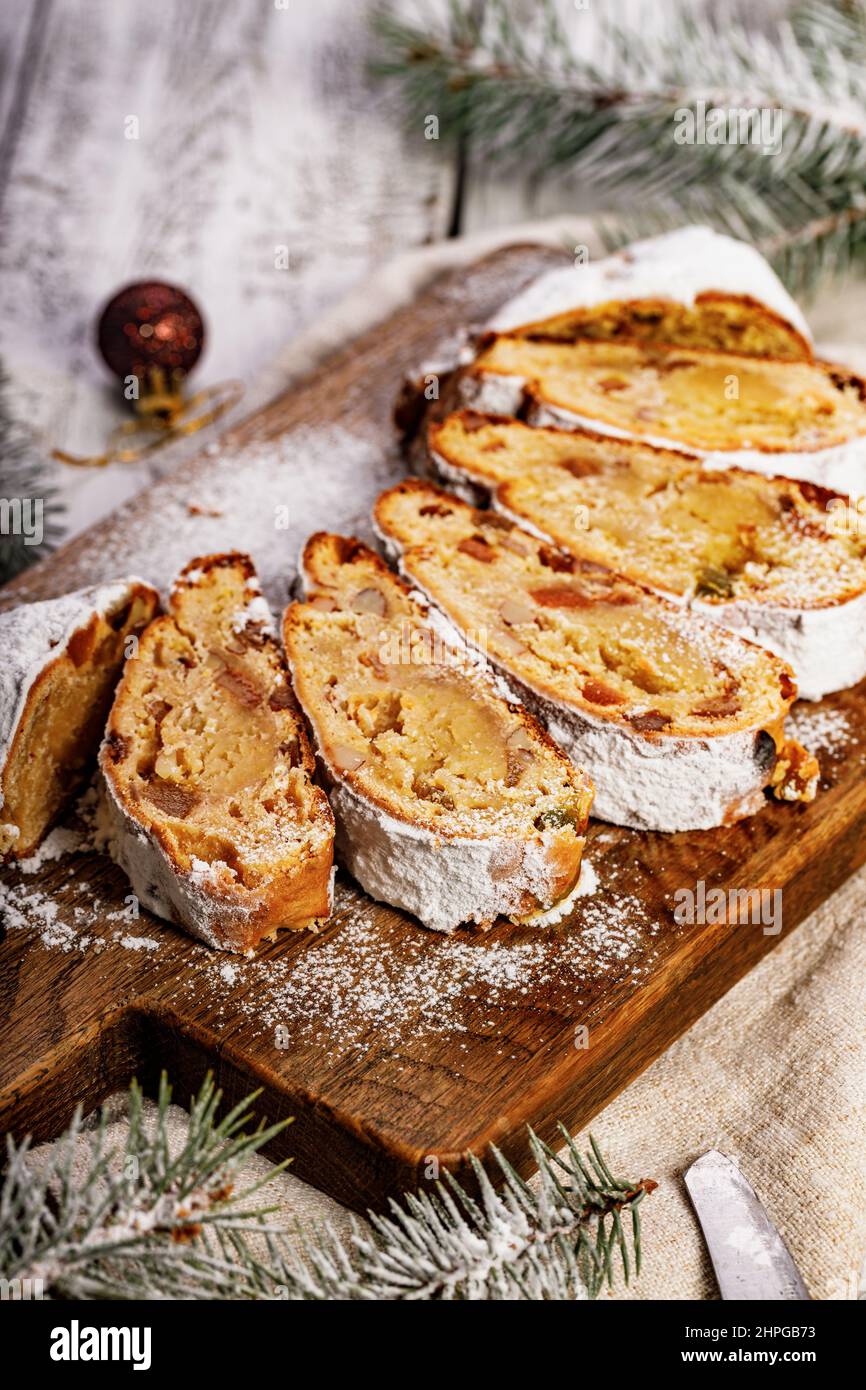 Stollen di Natale su un tagliere. Sfondo bianco tavolo in legno. Tradizionale dolce natalizio con marzapane, noci, uvetta e frutta secca. Verticale Foto Stock