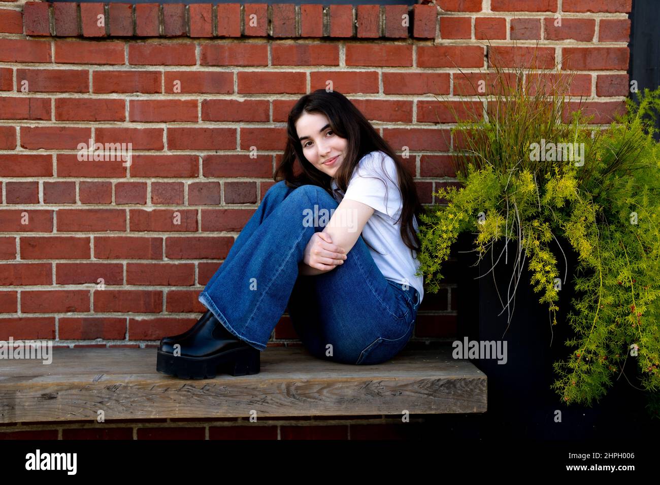 Giovane donna seduta su una panca di legno di fronte al muro di mattoni Foto Stock