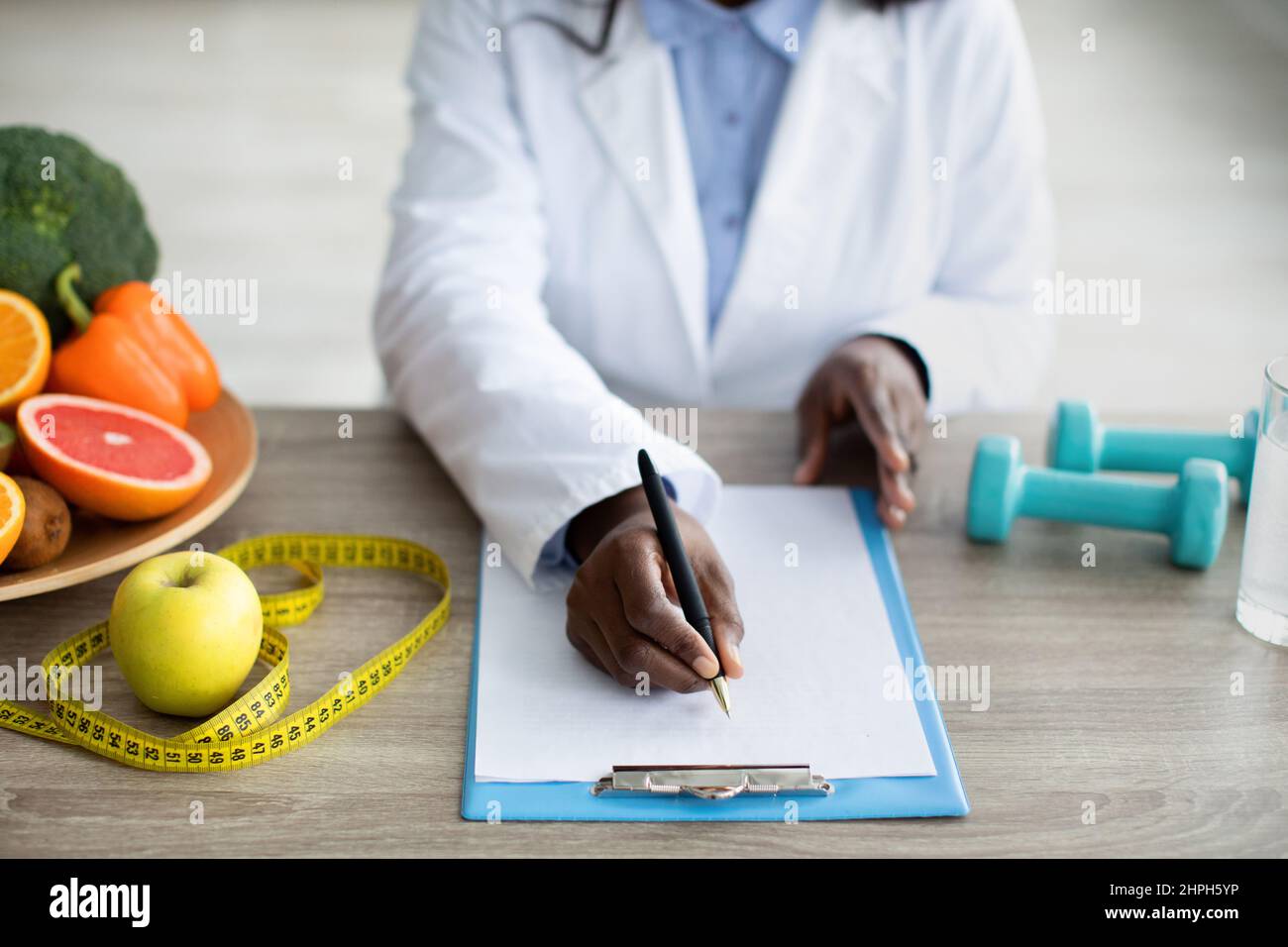 Vista croppata del pasto di pianificazione femminile nero per il cliente, scrittura negli appunti, seduta alla scrivania della clinica Foto Stock