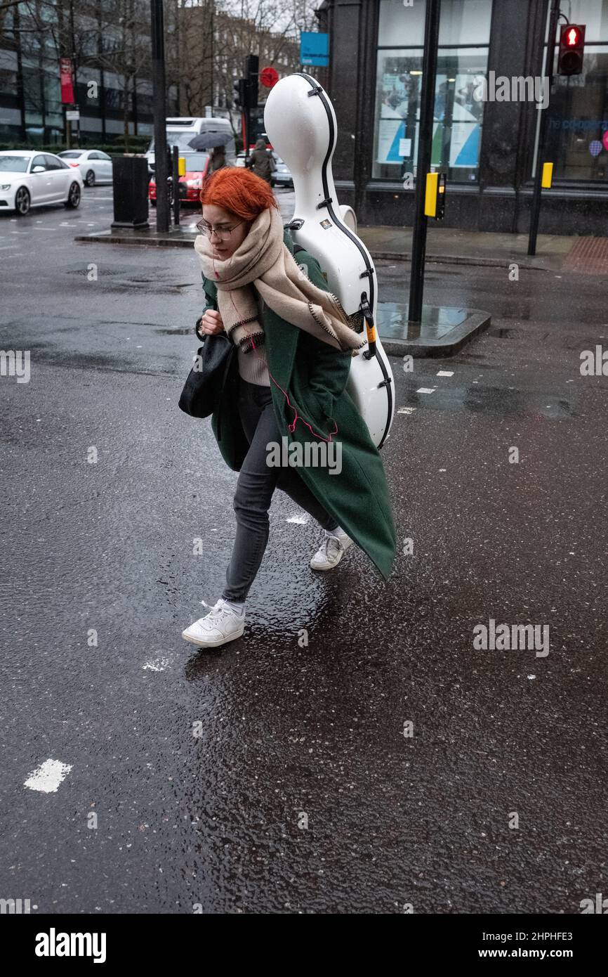 La femmina attraversa la strada a Londra durante una doccia a pioggia portando un grande strumento musicale in una custodia appositamente costruita Foto Stock