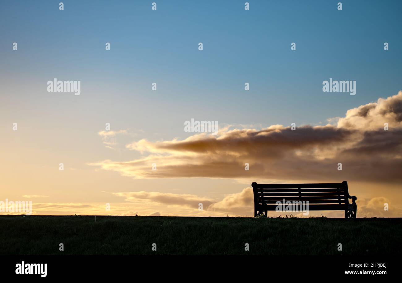 Una panchina a sagome al mare, presa nel tardo pomeriggio, con il cielo che diventa dorato Foto Stock