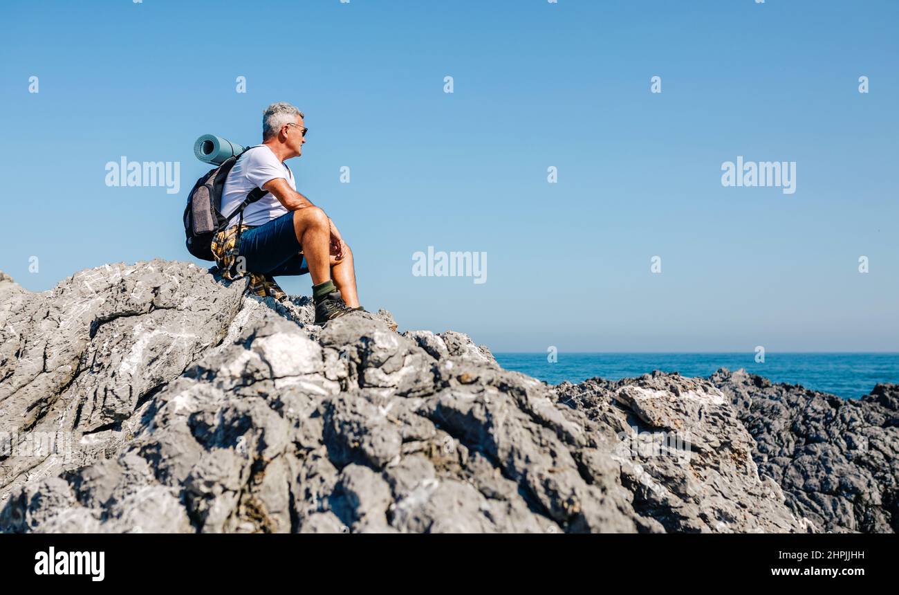 Uomo anziano escursioni guardando il paesaggio marino seduto sulle rocce Foto Stock