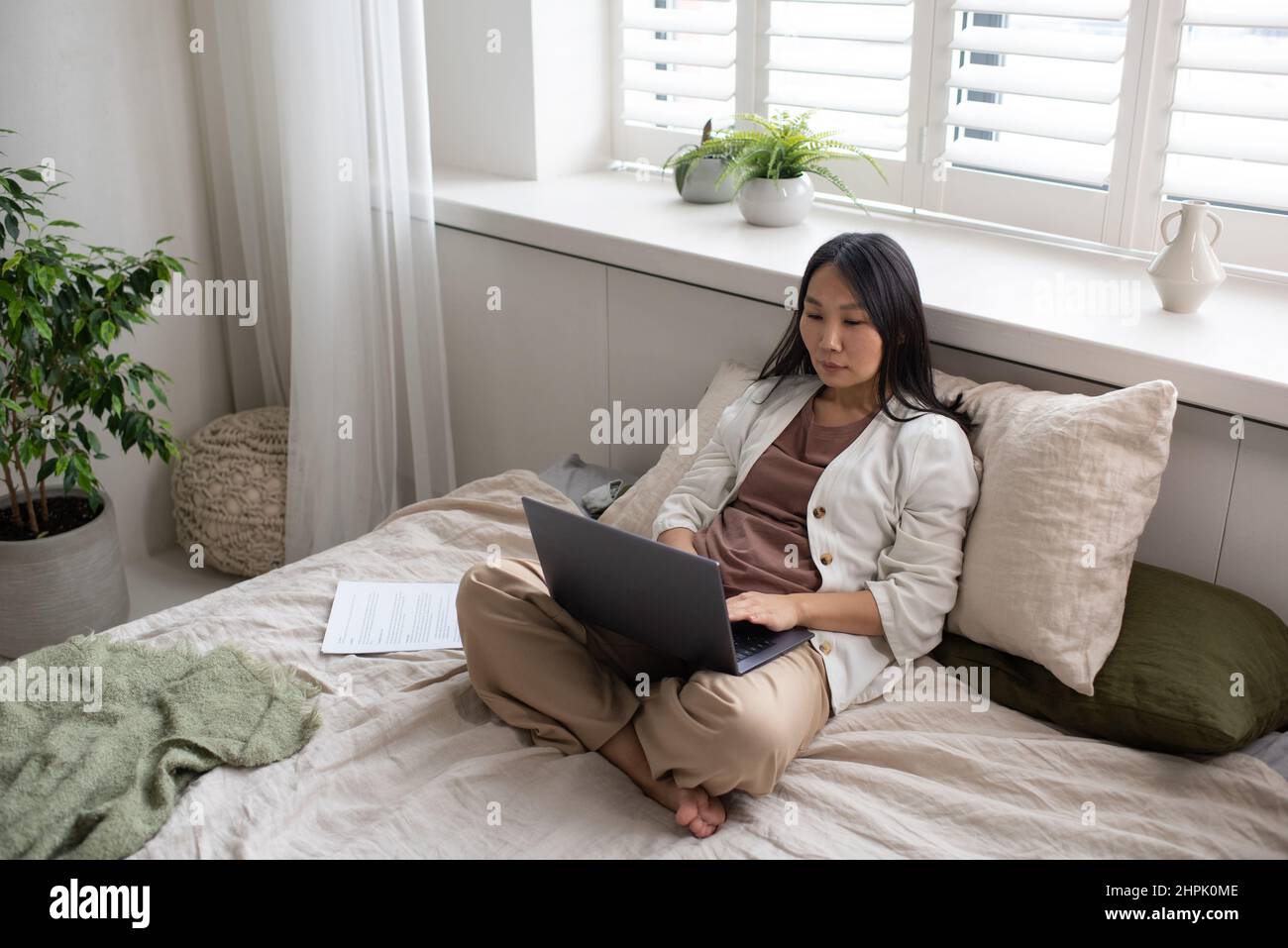 Giovane donna d'affari seria in casualwear digitando sulla tastiera del laptop mentre si siede sul letto matrimoniale in camera da letto durante il lavoro in ufficio domestico Foto Stock