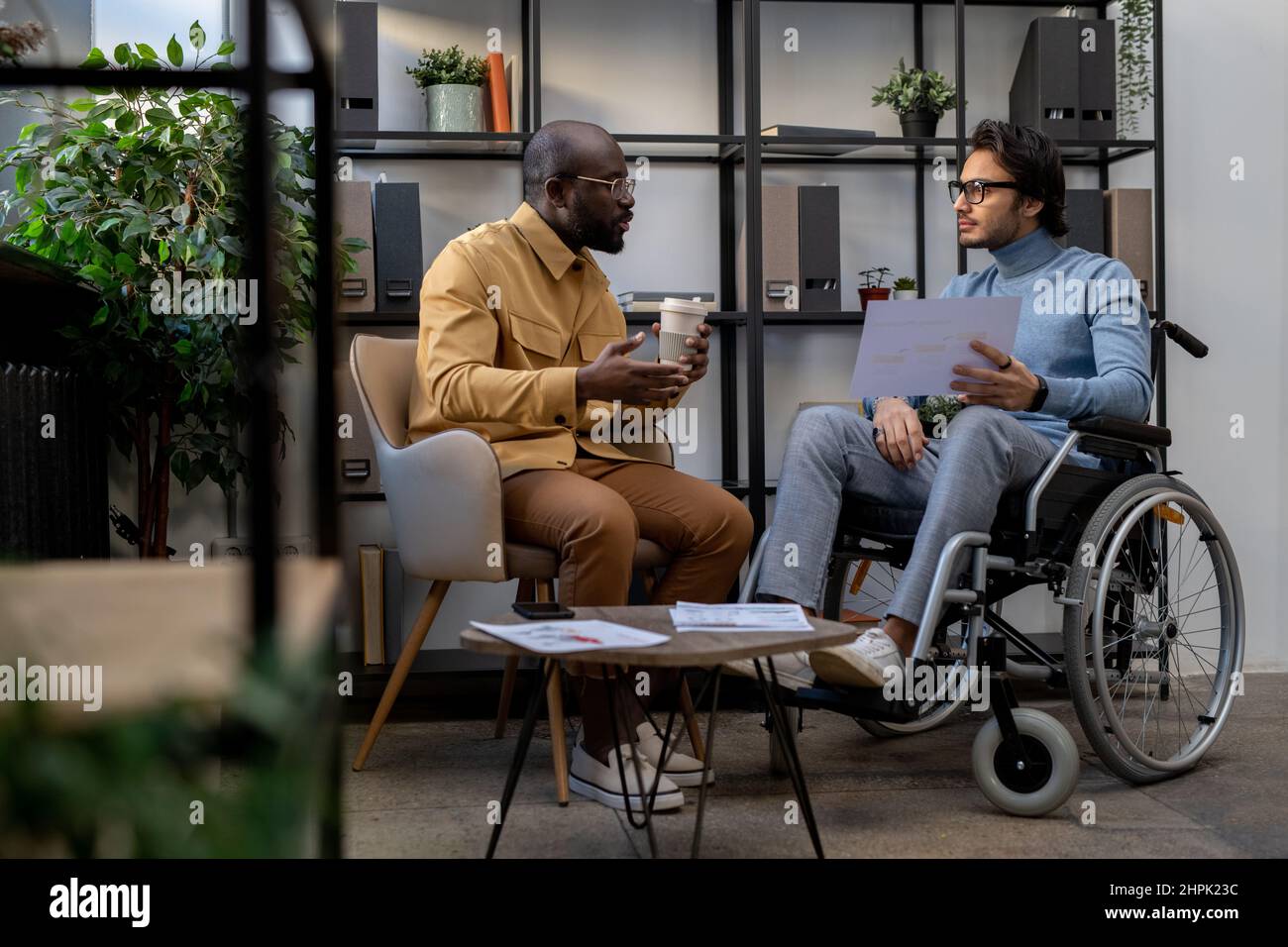 Uomo d'affari afroamericano con bevanda che comunica con il suo collega caucasico in sedia a rotelle alla riunione di lavoro in ufficio urbano Foto Stock