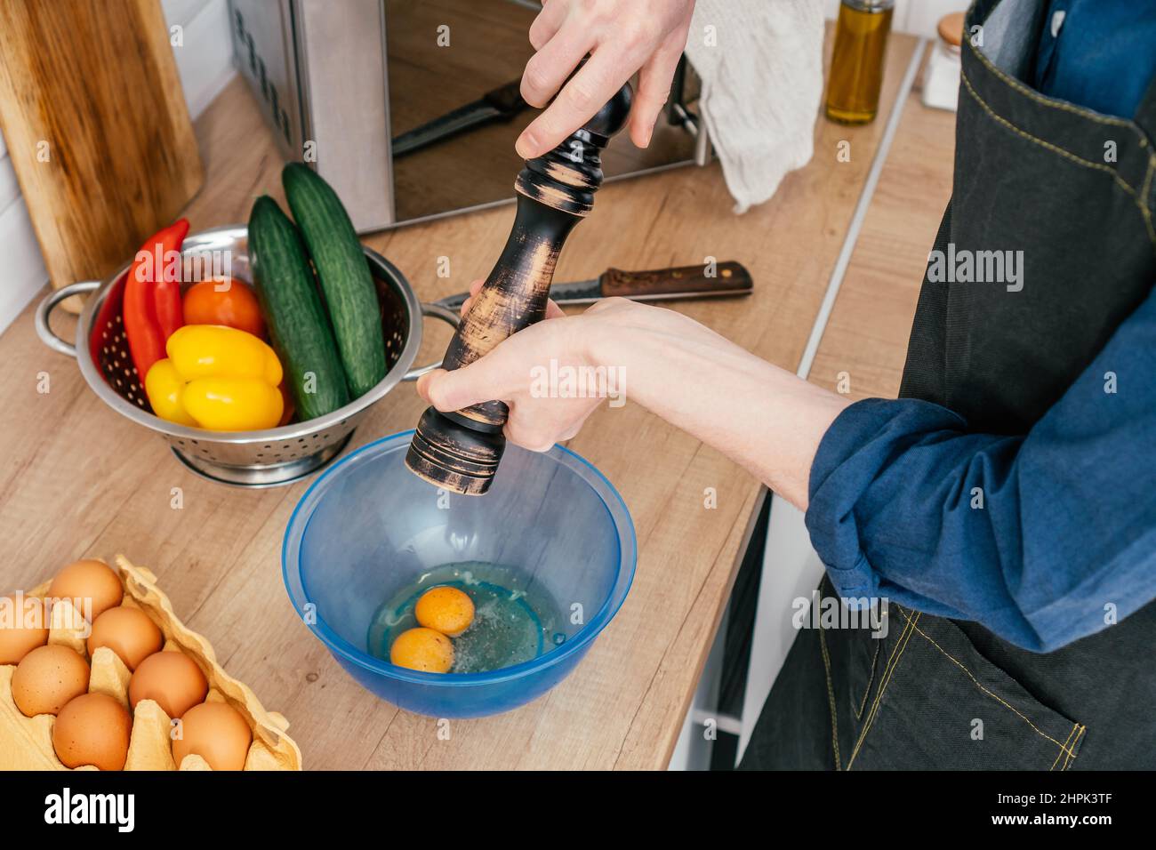 Vista dall'alto di un uomo irriconoscibile che pepperisce due uova con macinacaffè di legno in ciotola di plastica blu sul tavolo vicino al colino d'acciaio con campana rossa e gialla pe Foto Stock