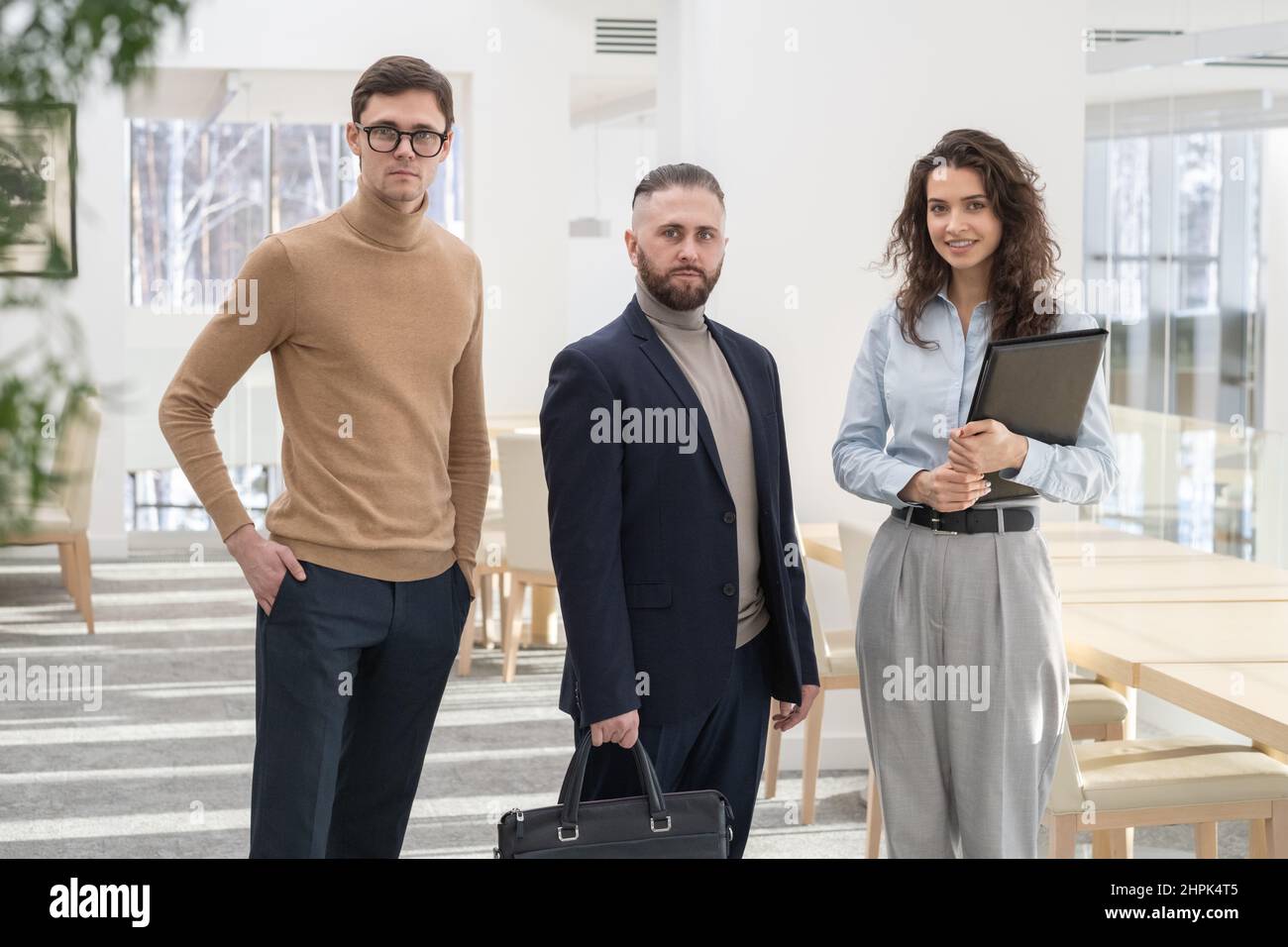 Gruppo di tre giovani collaboratori o partner aziendali di successo in abbigliamento formale davanti alla telecamera dopo un incontro di lavoro o una negoziazione Foto Stock
