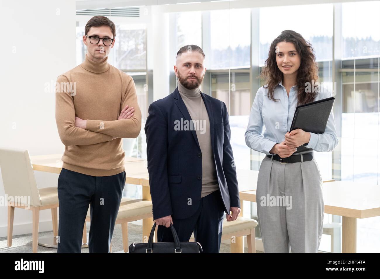 Datore di lavoro di mezza età di Cofident in tuta in piedi tra due giovani subordinati maschili e femminili in abbigliamento formale contro l'interno dell'ufficio moderno Foto Stock