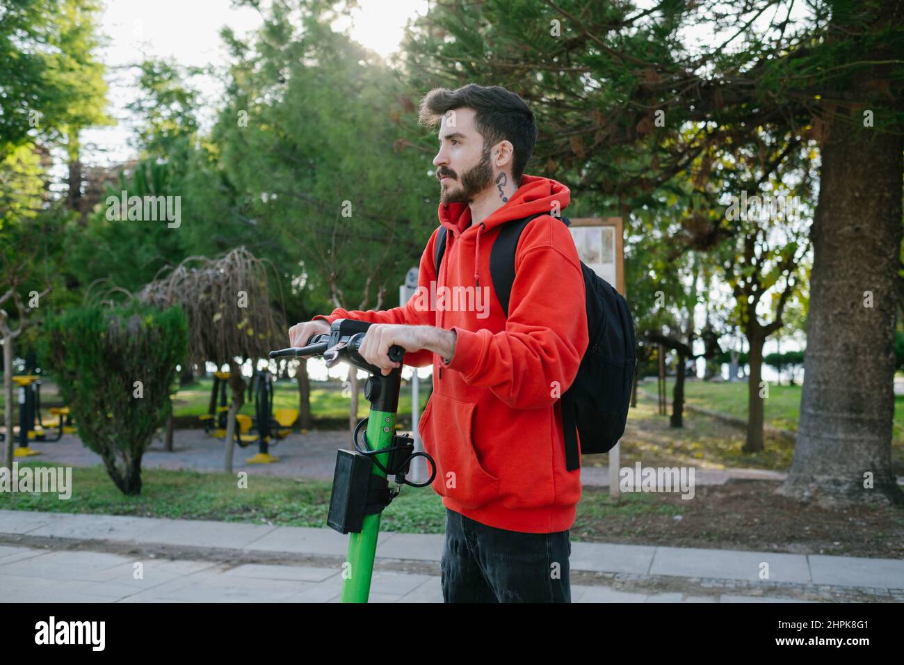 Guida scooter elettrico, giovane e attraente studente con zaino sta guidando il suo scooter a luminoso parco di giorno di sole sfondo. Foto Stock