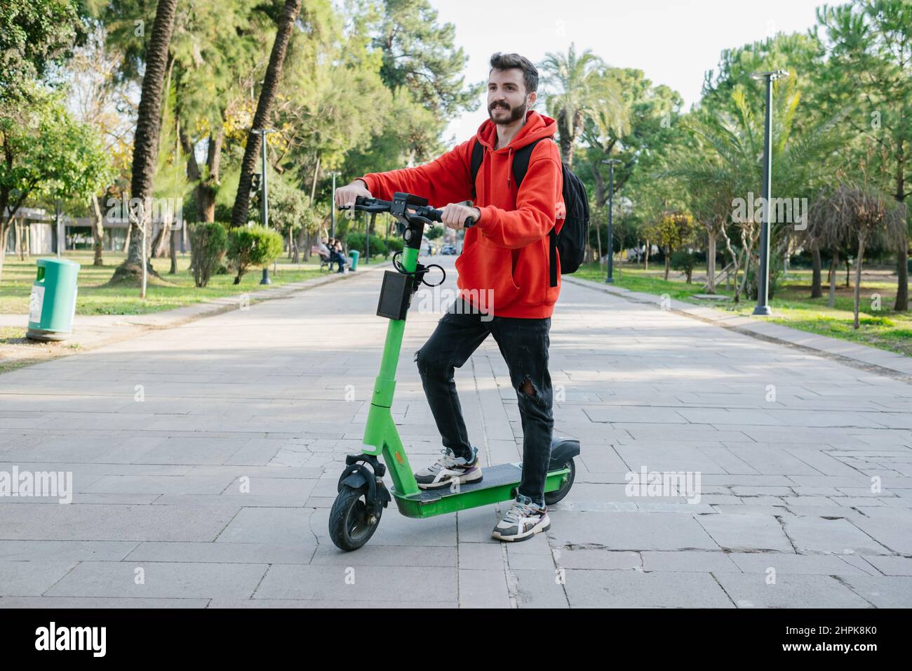 Uomo Hipster con scooter elettrico all'aperto nel parco. Giovane e attraente studente con zaino sta guidando il suo scooter a giorno di sole luminoso. Foto Stock