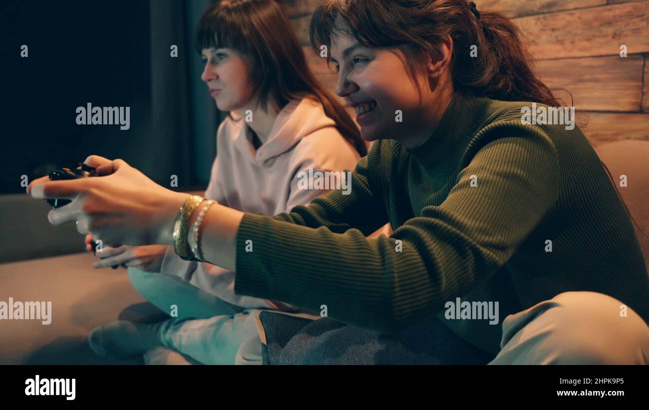 Due giovani donne che giocano a console videogioco con controller gamepad seduti sul divano. Videogamer di sesso femminile che si divertono e sorridono a casa insieme all'intrattenimento online. Foto Stock
