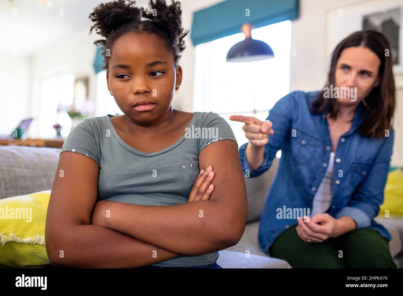 Madre caucasica matura che punta mentre spolding figlia afroamericana seduta con le braccia incrociate Foto Stock