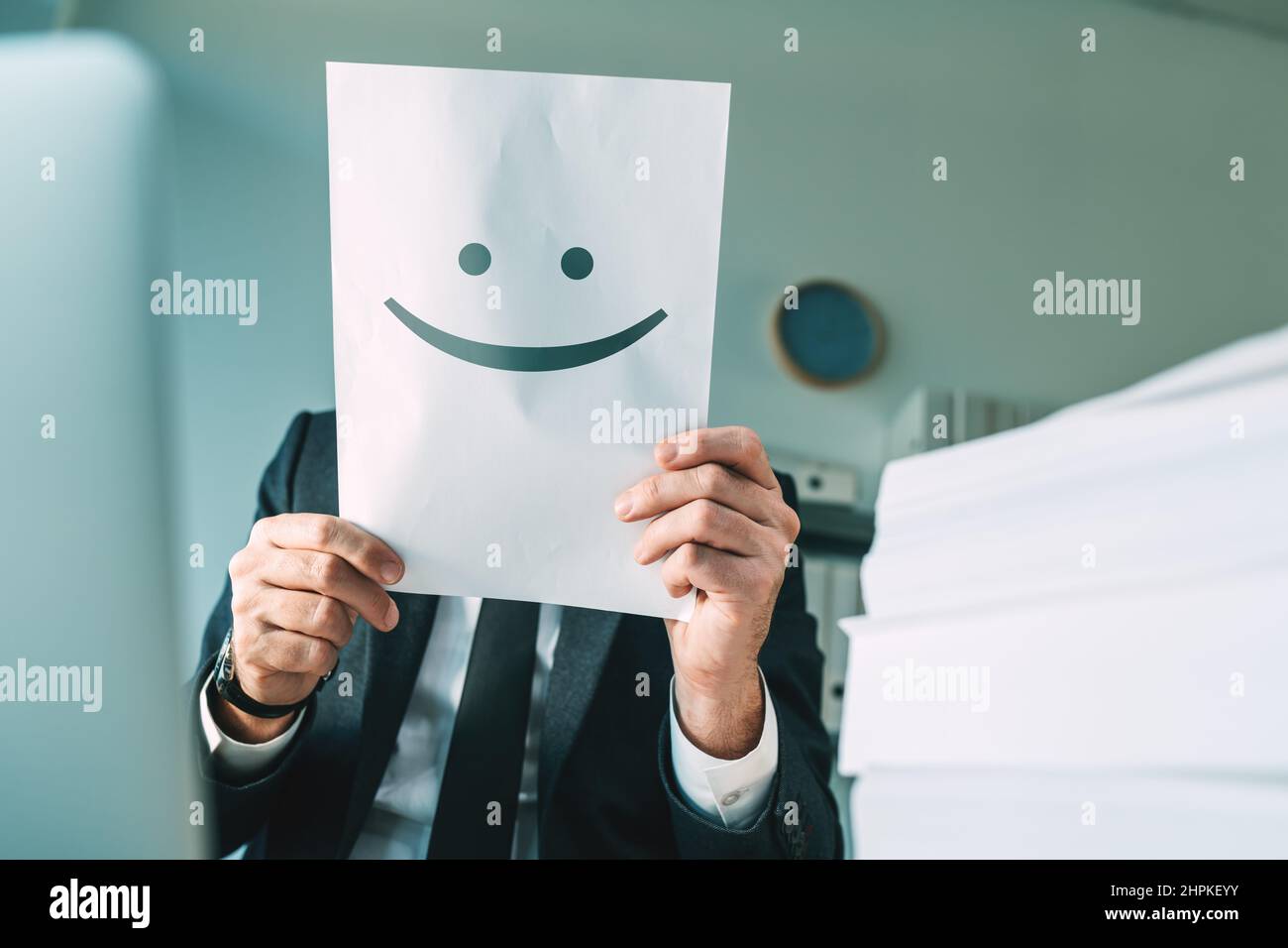 Felice uomo d'affari che tiene carta con emoticon sorridente in ufficio interno, fuoco selettivo Foto Stock