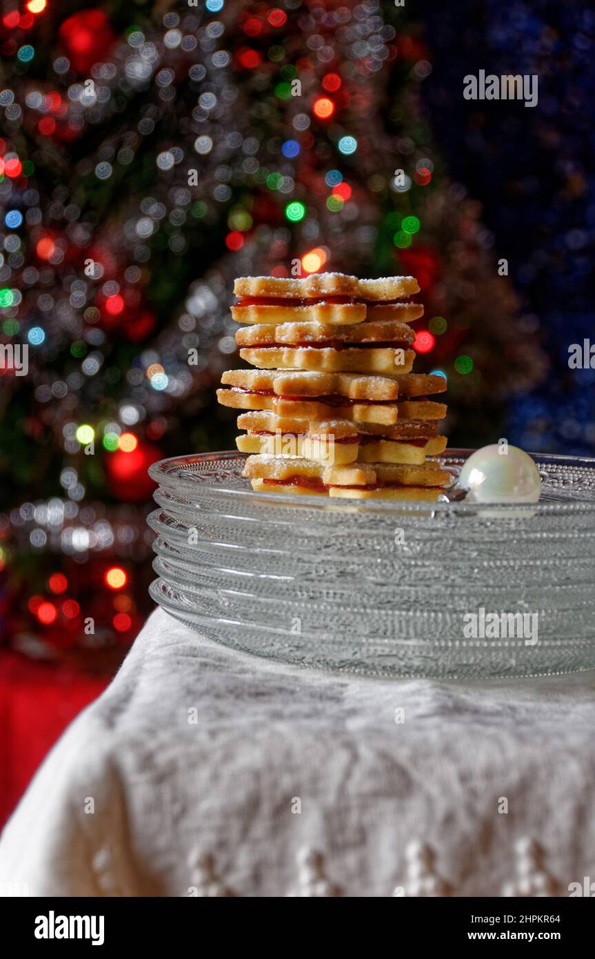 Biscotti fatti in casa con marmellata e zucchero a velo, serviti in piccole piastre di vetro su tessuto bianco, albero di natale in background con luci accese Foto Stock
