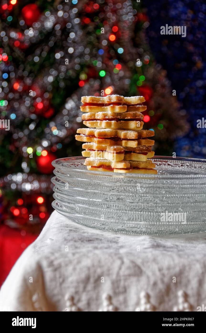 Biscotti fatti in casa con marmellata e zucchero a velo, serviti in piccole piastre di vetro su tessuto bianco, albero di natale in background con luci accese Foto Stock