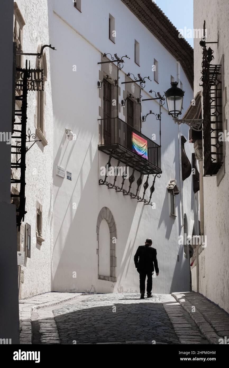 L'uomo cammina su una strada stretta a Sitges, Spagna. Vista posteriore. La luce del sole colpisce la facciata dell'edificio. Bandiera della comunità LGBT appesa su una terrazza. Foto Stock