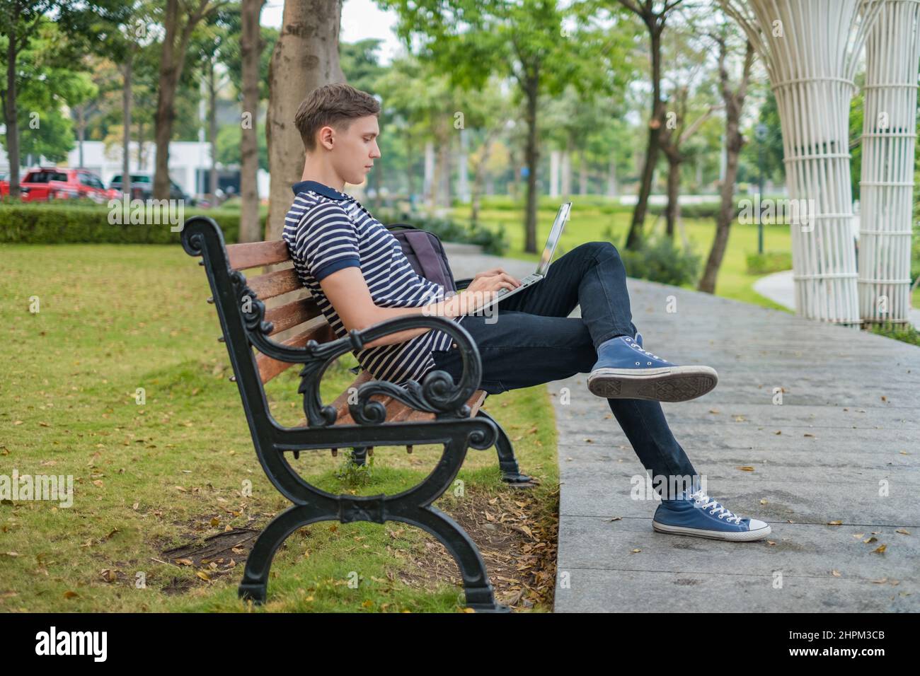Giovane uomo caucasico seduto sulla panchina del parco e lavorando sul computer portatile. Studente digitando su computer, freelance, lavorando online. Alta qualità Foto Stock
