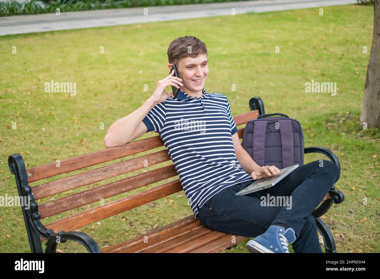 Giovane uomo caucasico seduto sul banco del parco, lavorando sul portatile e parlando al telefono, freelance. Studente che effettua una telefonata e sorride. Foto Stock