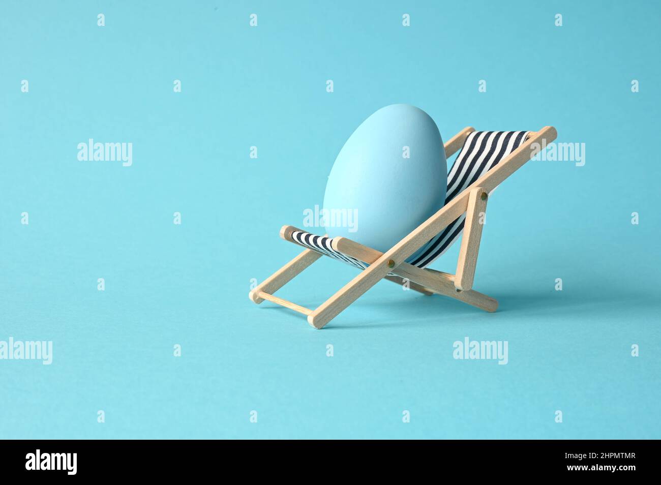 Composizione divertente e creativa con uovo di Pasqua seduto su sedia a sdraio su sfondo blu pastello. Minimo primavera o estate vacanza, vacanza e trave Foto Stock