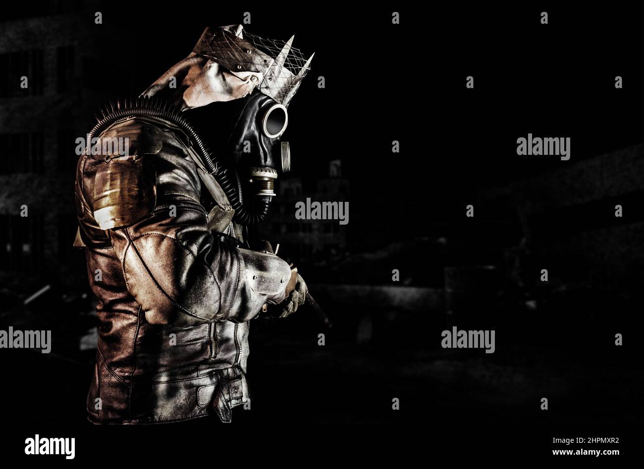 Foto del guerriero post-apocalittico con giacca blindata e corona di rottami vista laterale in piedi con fucile su sfondo scuro distrutta città. Foto Stock