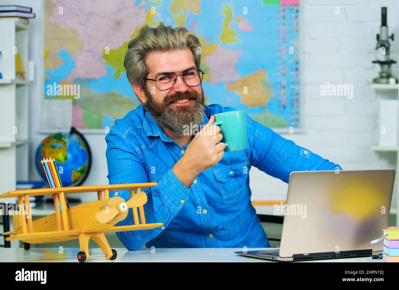 Insegnante sorridente con tazza di caffè in classe. Professore universitario seduto alla scrivania con computer portatile. Foto Stock