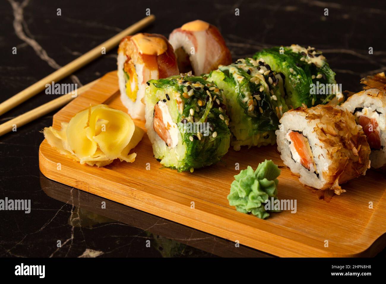 Rotoli di sushi freschi e gustosi di tipi diversi giacciono su un vassoio di legno su sfondo scuro Foto Stock