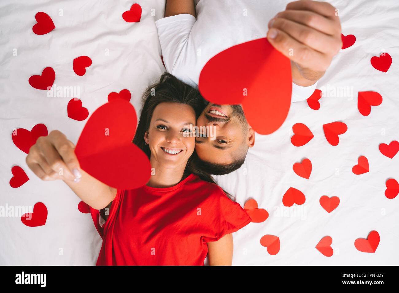 Vista dall'alto di una giovane coppia felice in amore che tiene i cuori di carta rossa mentre si trova sul letto bianco a casa Foto Stock