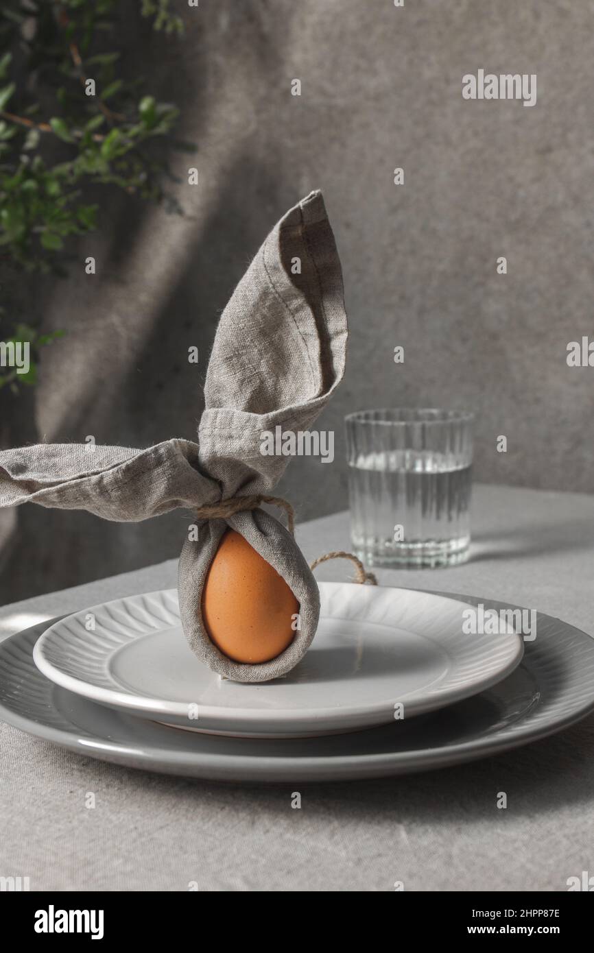 Festa Pasqua impostazione tavola. Divertente coniglietto pasquale fatto di uovo e tovagliolo di lino sul piatto. Sfondo grigio. Foto Stock