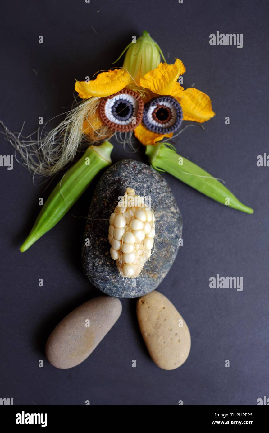 Faccia divertente allestito da cibo, prodotto agricolo da giardino sul tetto, mais piccolo, fiore di zucca, seta di mais, okra frutta, ciottoli e occhi uncinetto da Foto Stock