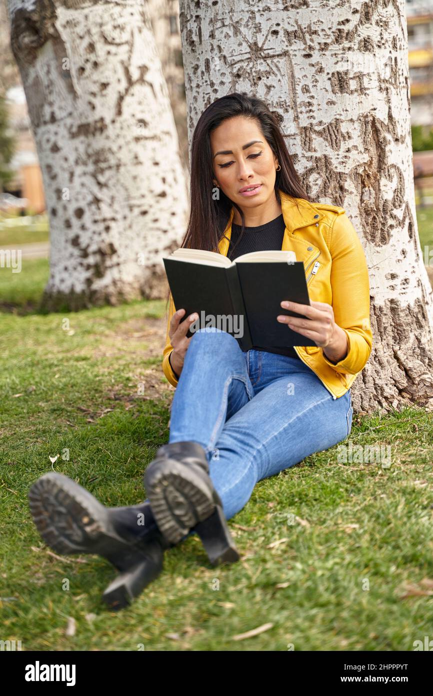 Pensierosa femmina ispanica seduta su erba sotto l'albero e lettura interessante storia a libro mentre godendo il fine settimana in giardino Foto Stock