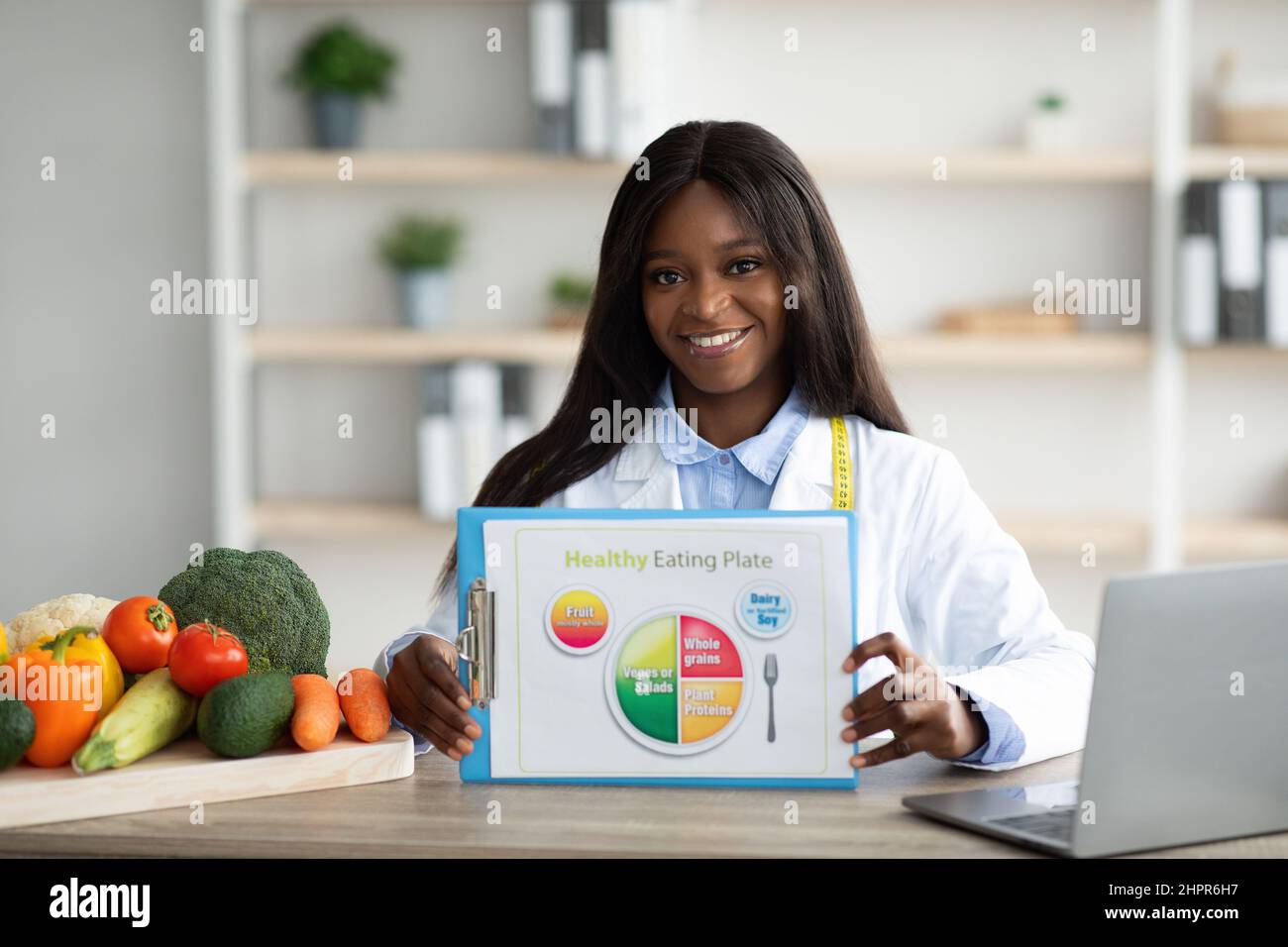 Buon dietista nero che mostra schematico sano mangiare piatto per la dieta, seduto sul posto di lavoro in clinica e sorridente Foto Stock