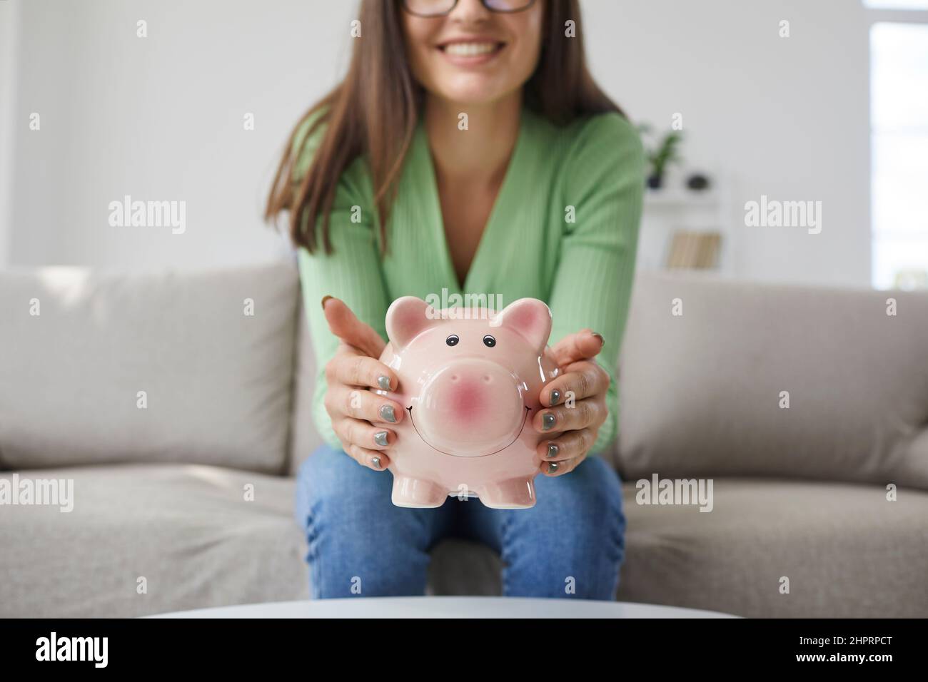 Primo piano di pig banca in forma di maiale rosa in mani di donna sorridente seduto sul divano a casa. Foto Stock