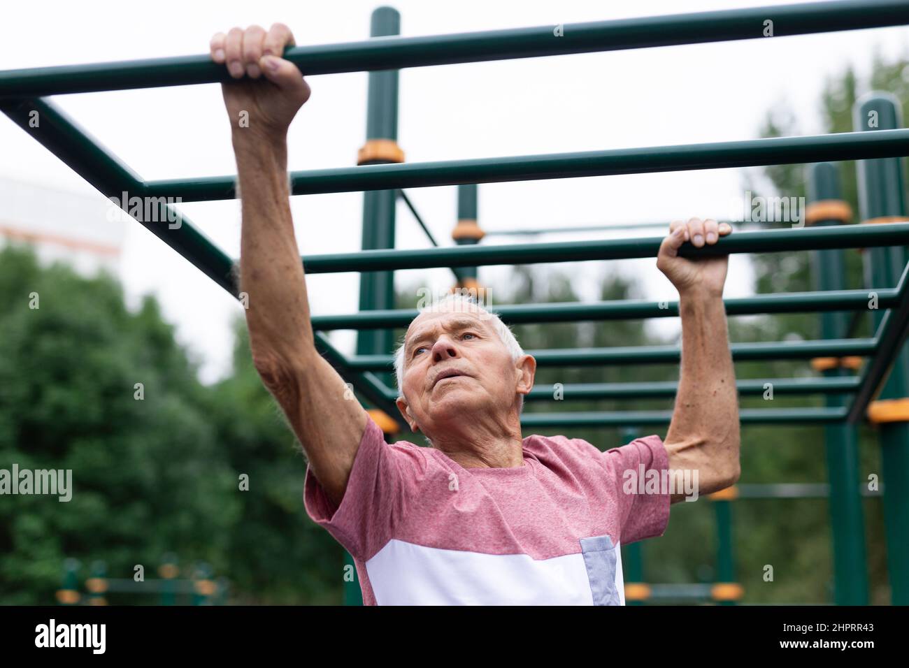 Il nonno attivo si muove con l'aiuto delle sue mani su una scala orizzontale sul terreno sportivo Foto Stock