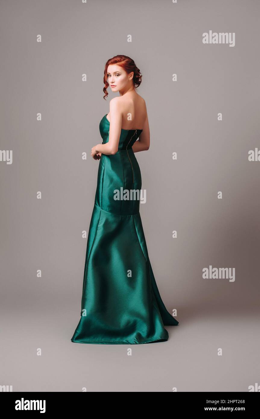 Abito da palla smeraldo. Giovane donna in abito da sera verde lungo. Donna zenzero in posa in studio. Bel modello con tacco alto, look femminile per una vigilia Foto Stock