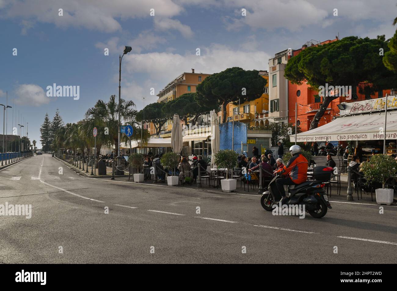 Uno scooter che passa sul lungomare di Sanremo con persone che pranzano in ristoranti all'aperto in una soleggiata giornata invernale, Imperia, Liguria, Italia Foto Stock