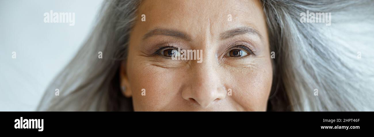 Donna asiatica di mezza età con capelli volanti hoary guarda in fotocamera su sfondo grigio chiaro Foto Stock