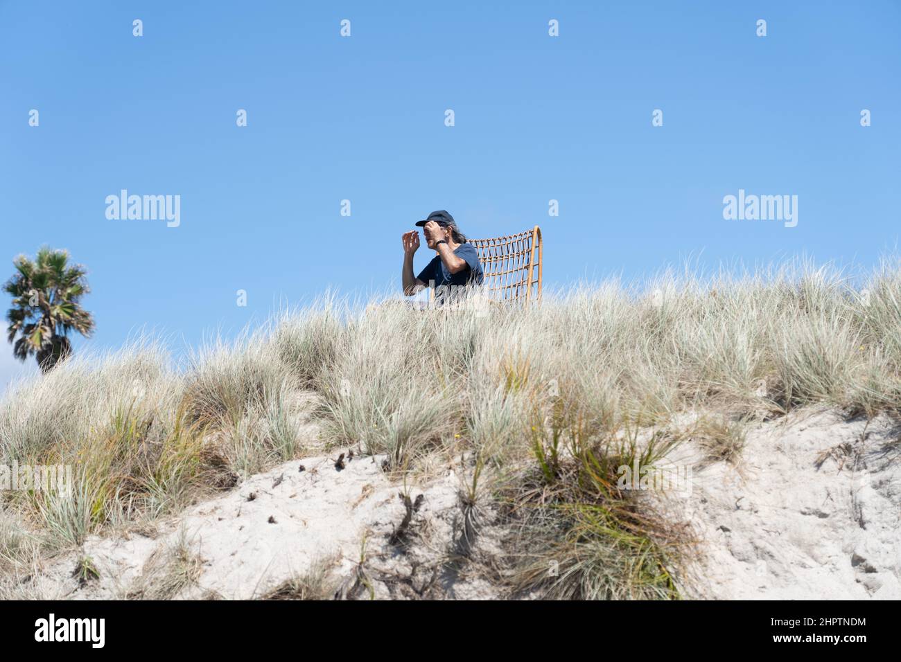Tauranga Nuova Zelanda - Febbraio 22 2022; uomo seduto in sedia sulle dune di sabbia di os superiore con le mani alzate proteggendo il sole dagli occhi mentre si guarda al mare Foto Stock