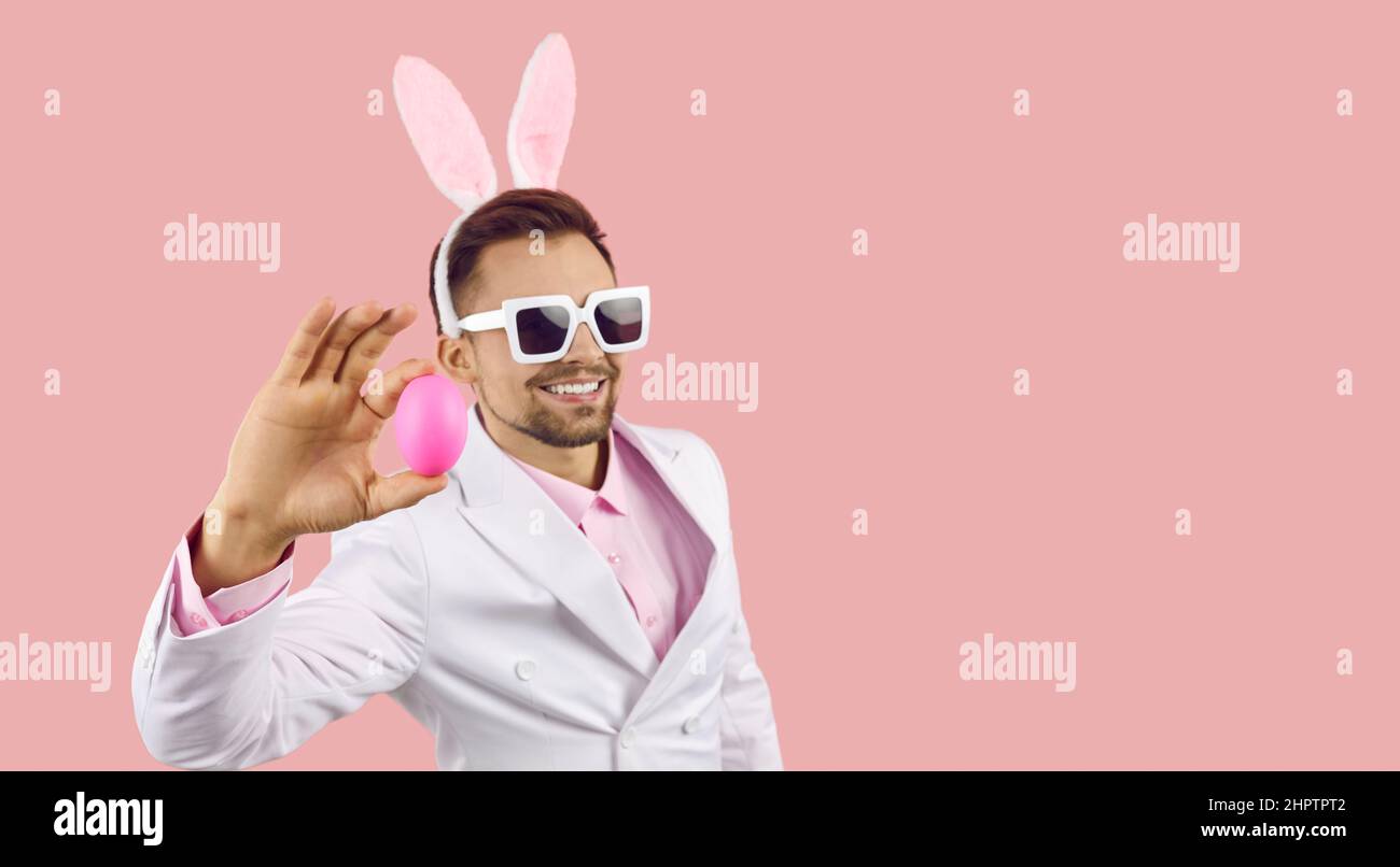 Bell'uomo in occhiali da sole e coniglietto orecchie che mostra l'uovo di Pasqua su sfondo rosa spazio copia Foto Stock