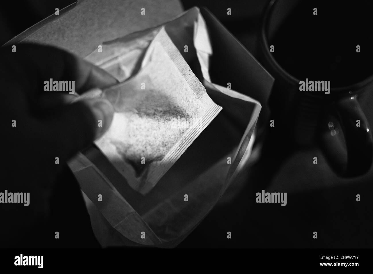 Una mano raggiunge per un sacchetto del tè da una scatola aperta vicino ad una tazza rossa su un tavolo di legno nella luce dell'ora d'oro. Immagine in bianco e nero. Foto Stock