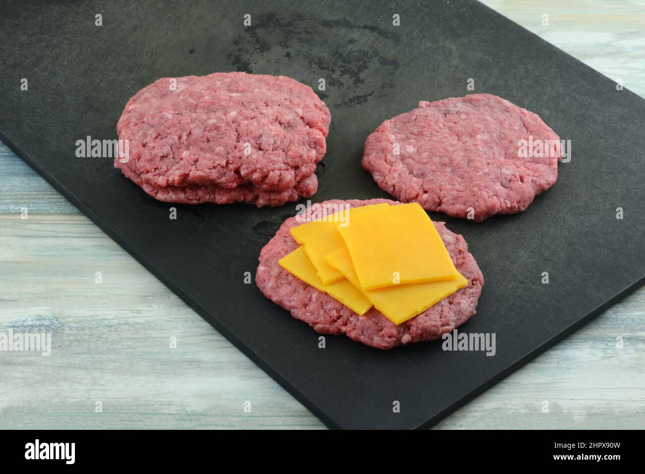 Disporre le fette di formaggio tra le polpettine di hamburger di manzo crudo macinato sul tagliere nero Foto Stock