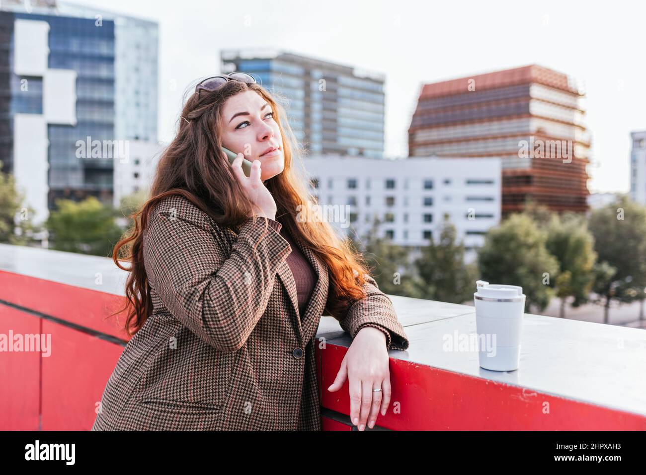 Donna pensiva in outerwear con capelli lunghi appoggiati sul bordo vicino al caffè da asporto e guardando via mentre si parla sullo smartphone sulla strada della città Foto Stock