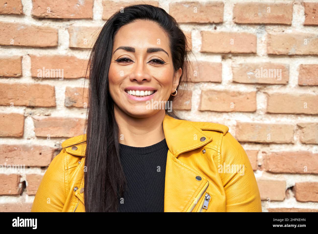 Donna ispanica ottimista con capelli scuri e giacca in pelle in piedi contro muro di mattoni in strada e guardando la macchina fotografica Foto Stock