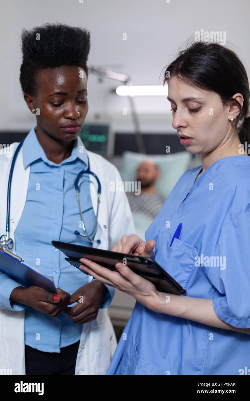 Infermiera che mostra i risultati di laboratorio da tablet digitale a medico che tiene appunti con storia medica nel reparto moderno dell'ospedale. Specialisti sanitari che utilizzano la tecnologia moderna per la diagnosi clinica. Foto Stock