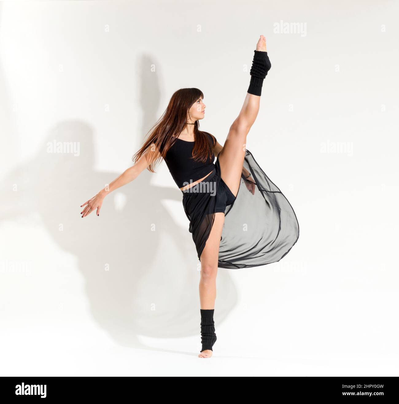 Intera lunghezza di ballerina femminile flessibile con lunghi capelli marroni in abiti neri che si sviluppano posa su sfondo bianco in studio Foto Stock