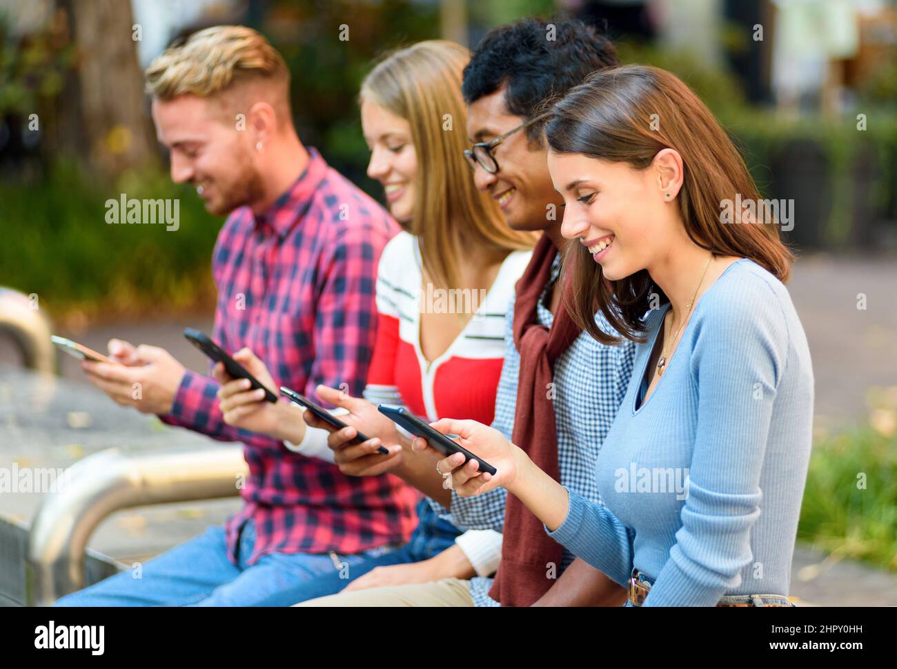 Gruppo di amici multirazziale positivi messaggi di testo su cellulari moderni mentre si siede insieme su strada con alberi verdi della città Foto Stock