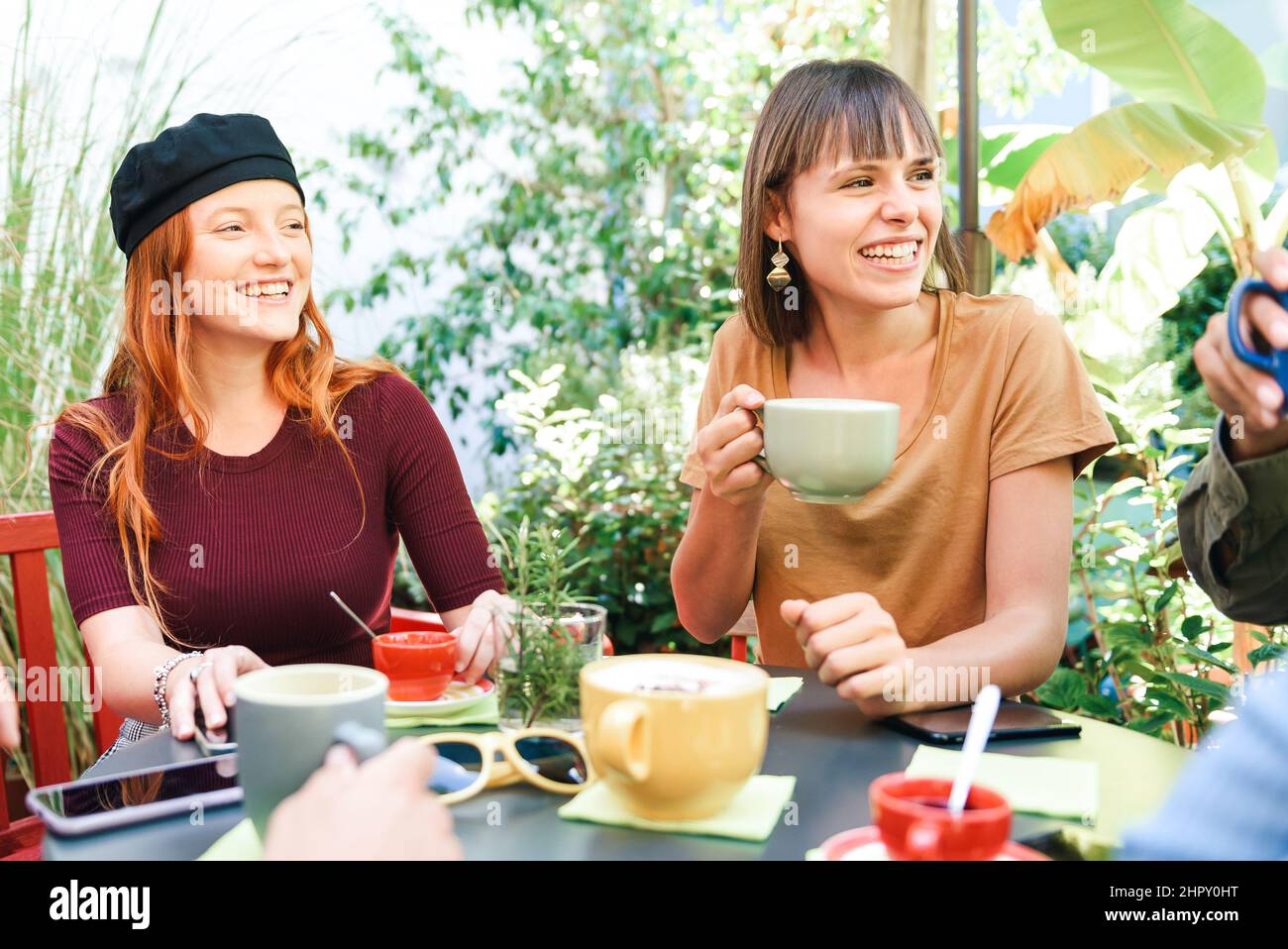 Gruppo di amici felici con tazze di caffè caldo seduti al tavolo in giardino all'aperto con alberi verdi durante la pausa Foto Stock
