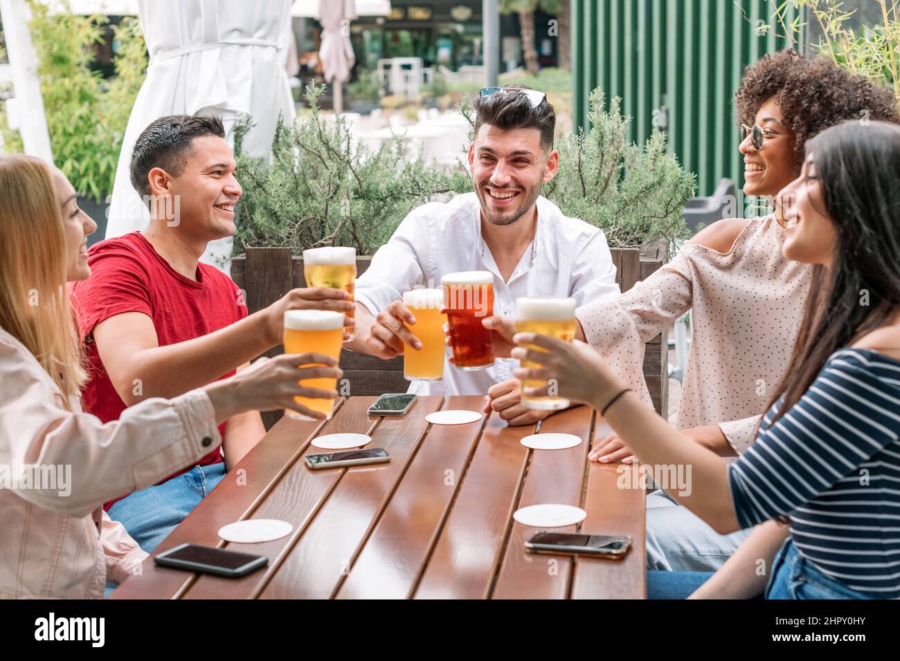 Compagnia di allegri giovani amici multirazziali tostare con bicchieri di birra mentre festeggia la riunione sulla terrazza del ristorante nel giorno d'estate Foto Stock